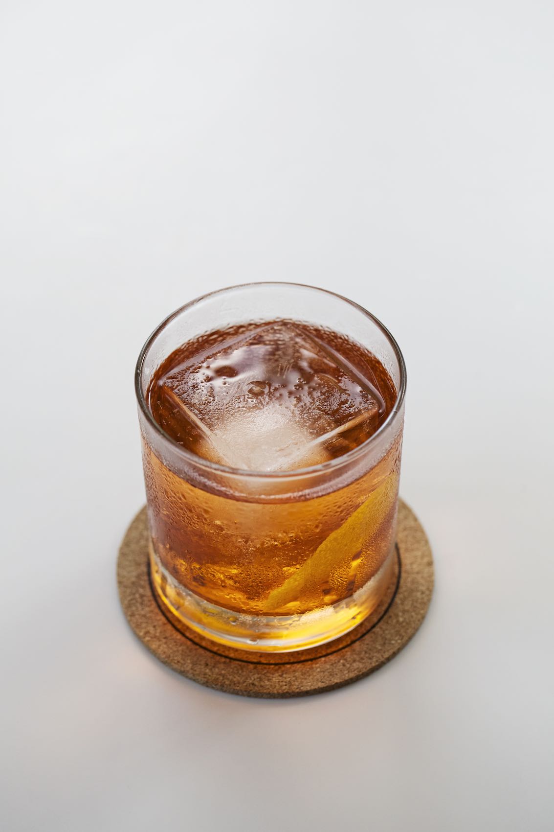 Jody Horton Photography - Whiskey with large cube on cork coaster on white table.