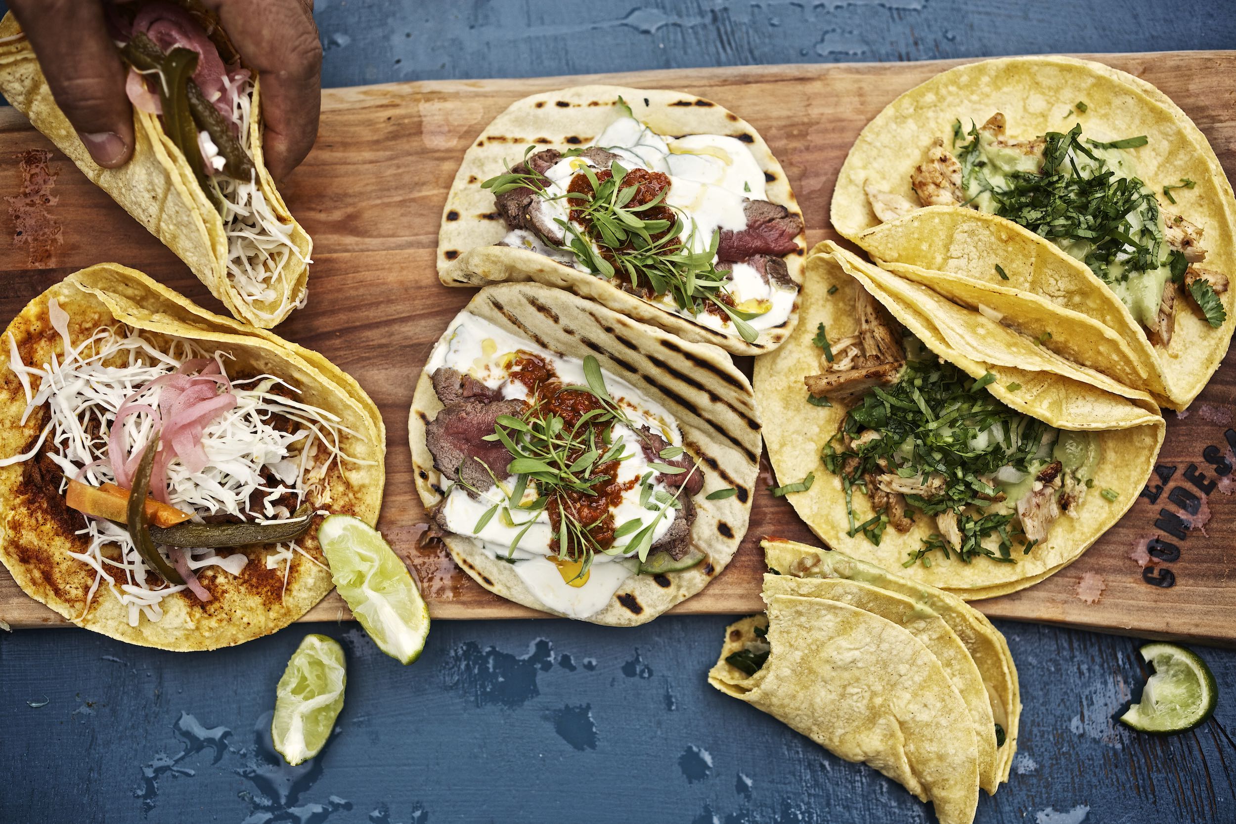 Jody Horton Photography - Assorted tacos on wood cutting board on blue table. 