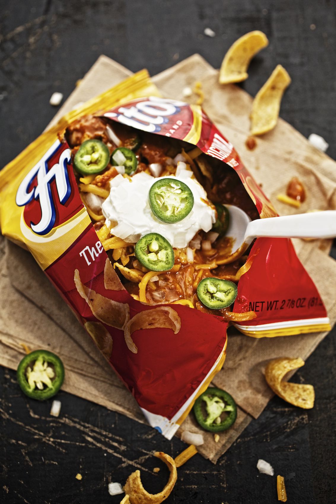 Jody Horton Photography - Frito pie served in Fritos bag with toppings, against black surface. 