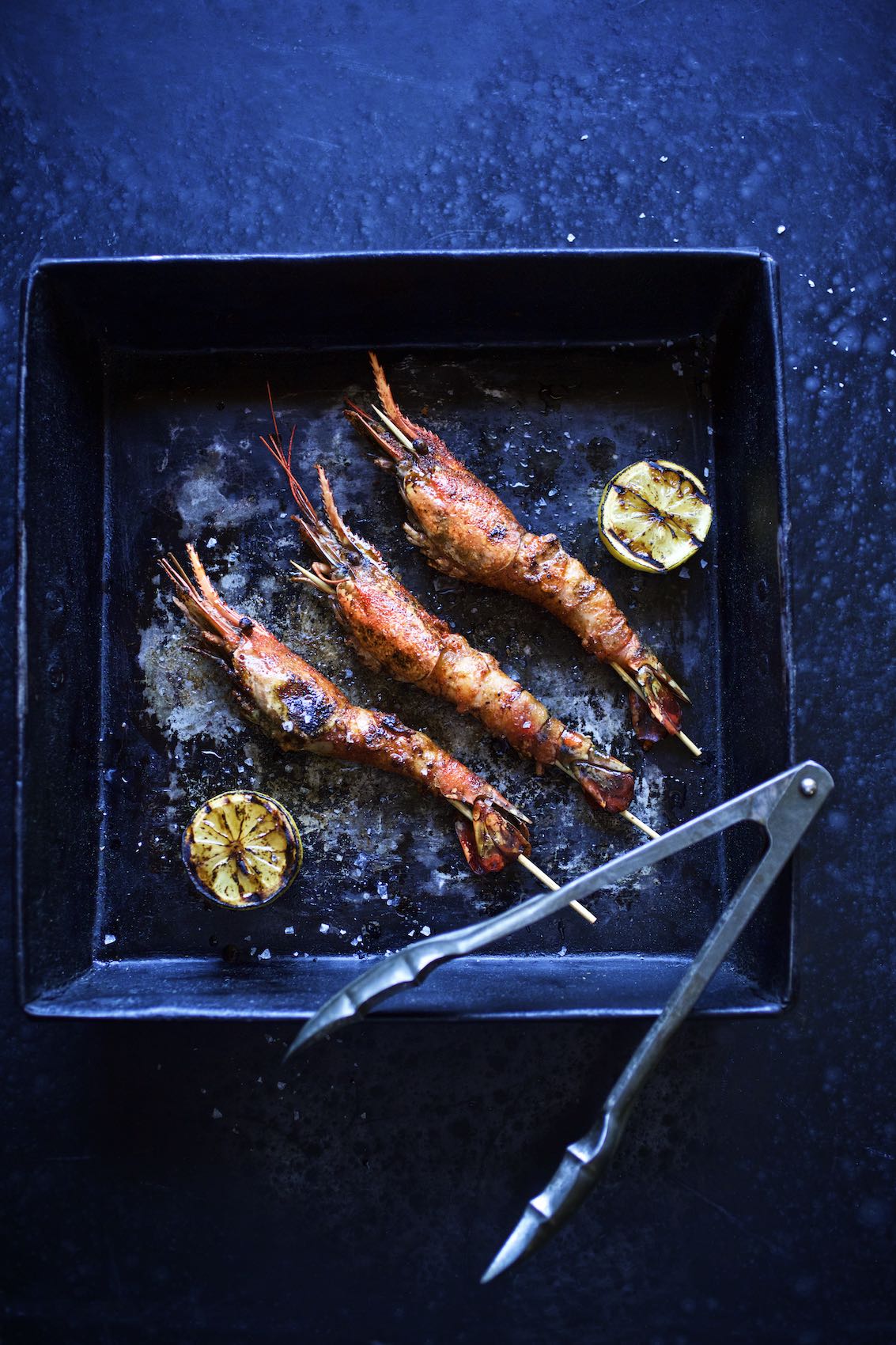 Jody Horton Photography - Skewered, head-on prawns and grilled lemon in rustic, black dish. 