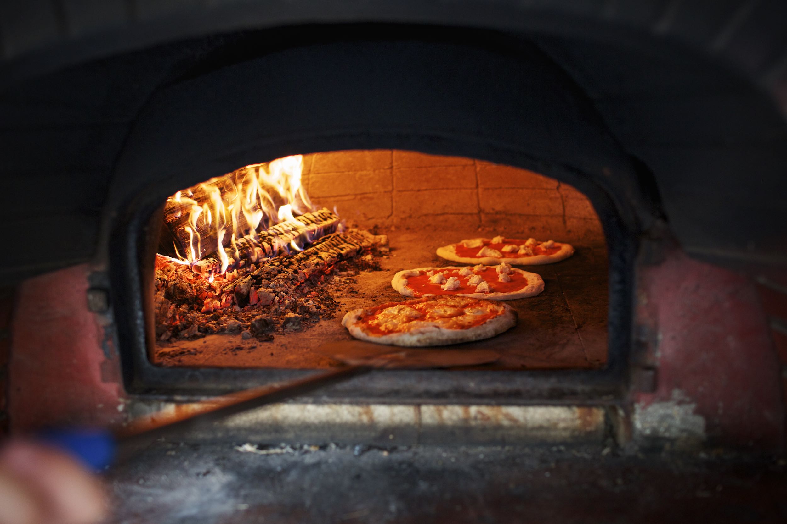 Jody Horton Photography - Three pizzas cooking in brick oven next to fire. 