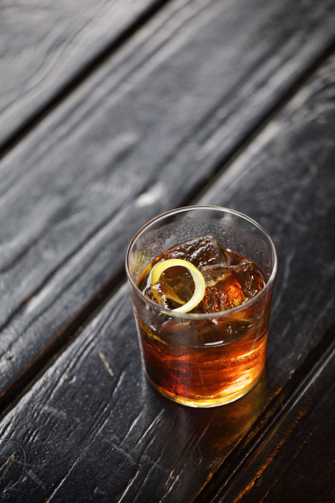 Jody Horton Photography - Iced bourbon cocktail with lemon twist on a dark, wood table. 