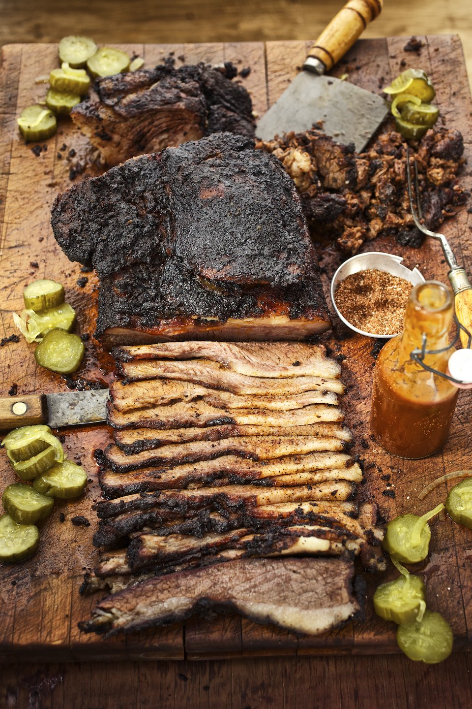 Half carved brisket with pickles and barbecue sauce on a wood cutting board. 