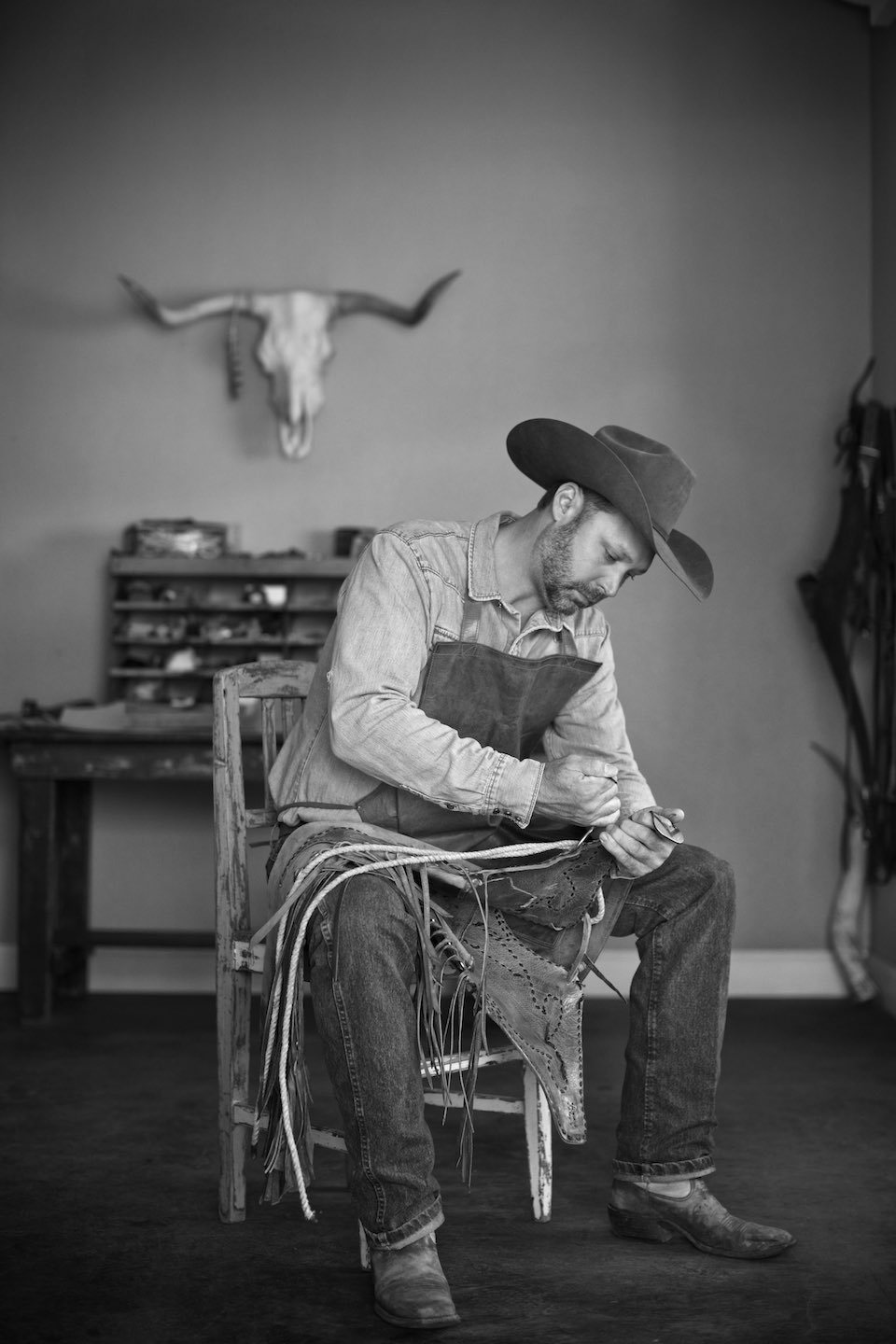 Leather worker sitting on a wood chair working on a piece, shot in B&W. 